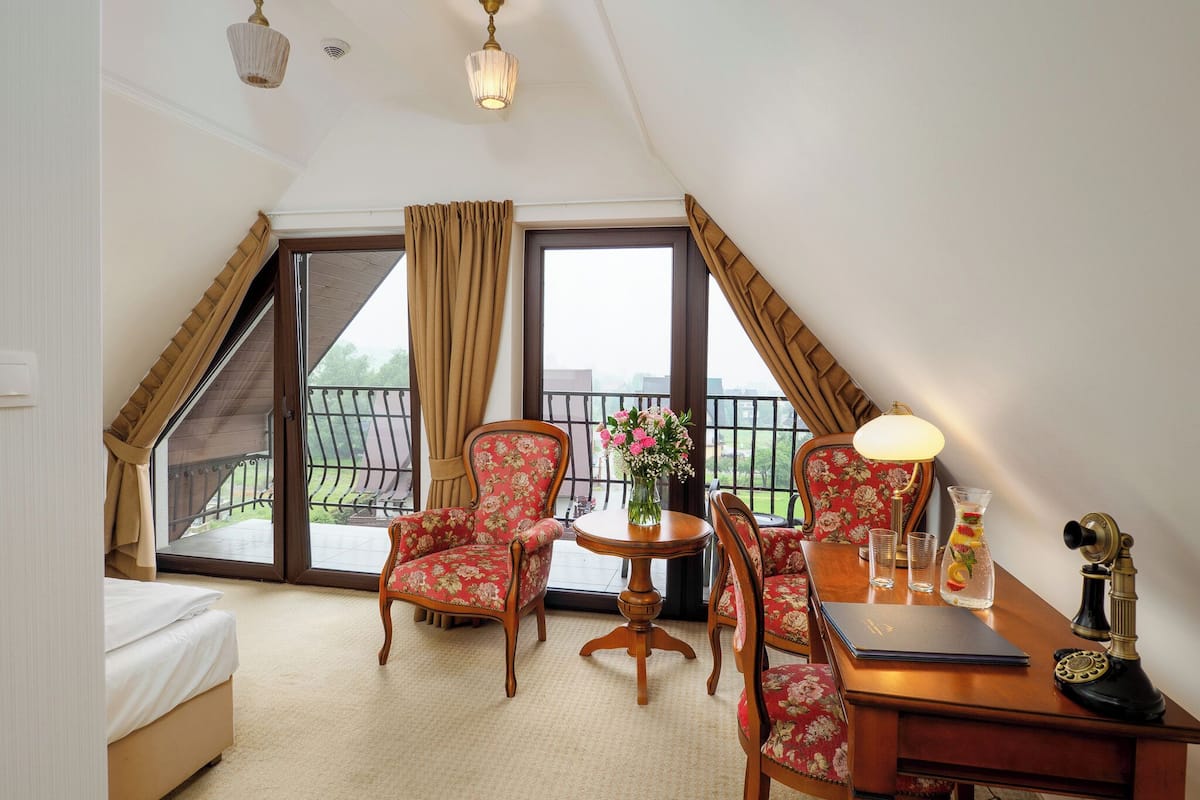 Hotelkamer met een bed, een bureau met lamp, telefoon, vaas met bloemen en een stoel. Er is een balkon met een hekwerk en uitzicht op de omgeving.
