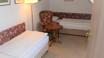 Um quarto pequeno e acolhedor com duas camas de solteiro, uma poltrona com padrão floral, uma mesa redonda de madeira e um abajur.