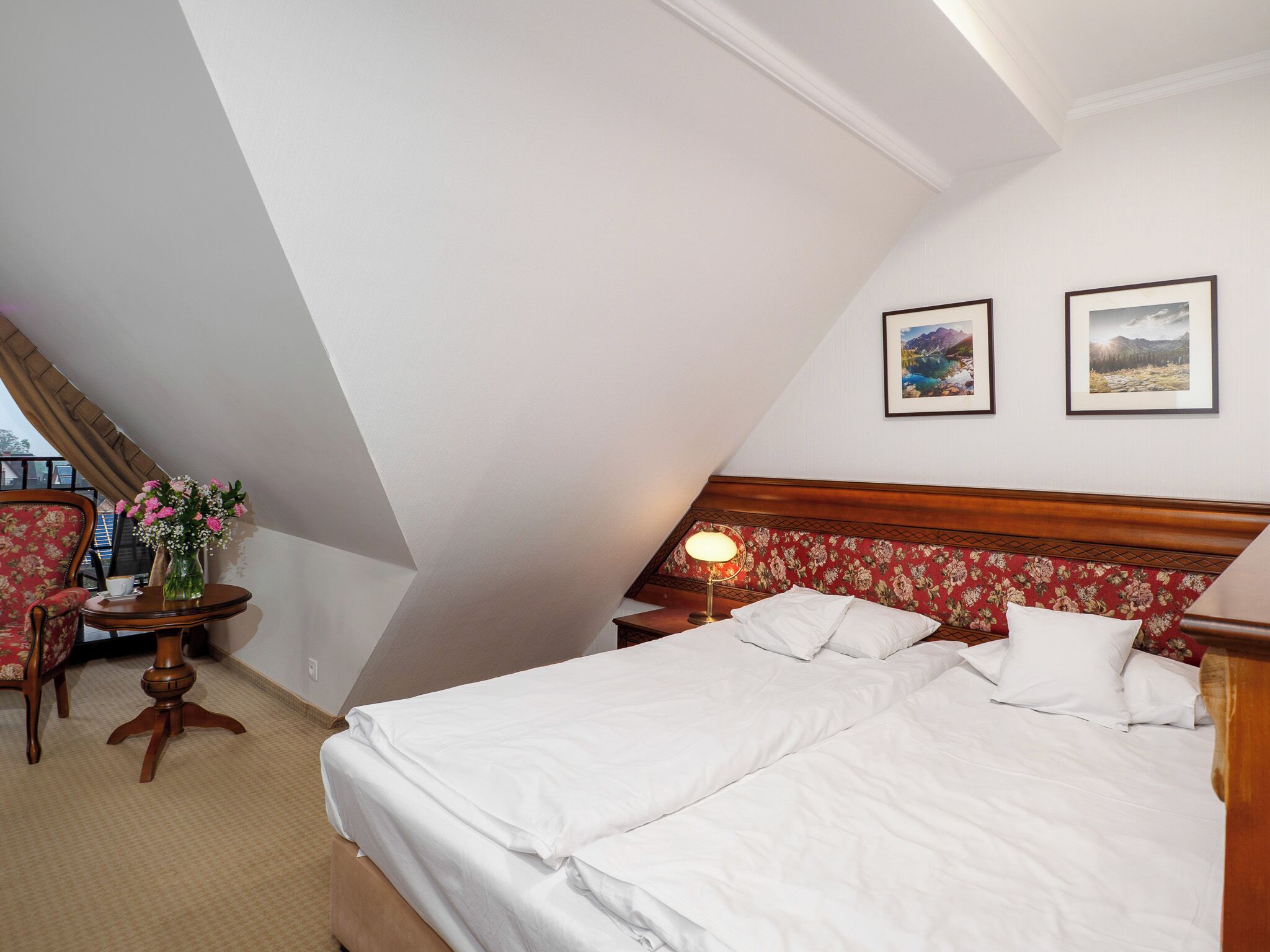A hotel room with a large bed, a wooden headboard, two framed pictures on the wall, a small table with a vase of flowers, and a chair.