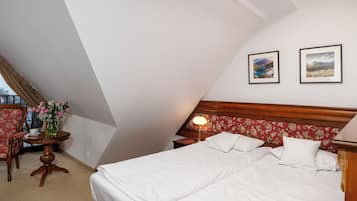A hotel room with a large bed, a wooden headboard, two framed pictures on the wall, a small table with a vase of flowers, and a chair.