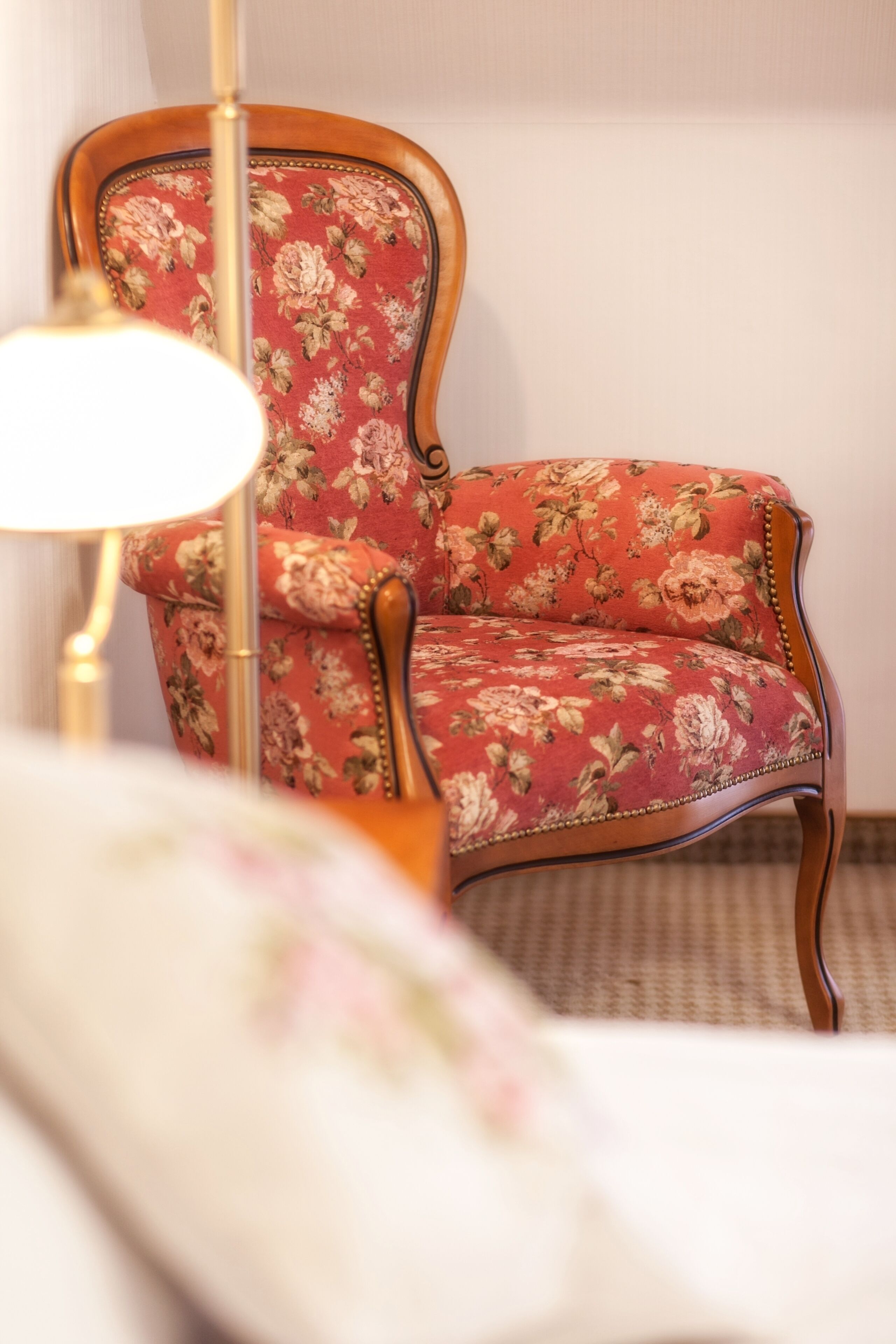 Een fauteuil met bloemenprint en houten frame, een vloerlamp en een bed met bloemenbedekking.