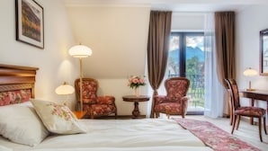 Une chambre d’hôtel avec un lit, deux fauteuils à motifs floraux, une petite table sur laquelle est posé un vase de fleurs, un bureau avec une lampe et une vue sur les montagnes par une grande fenêtre.