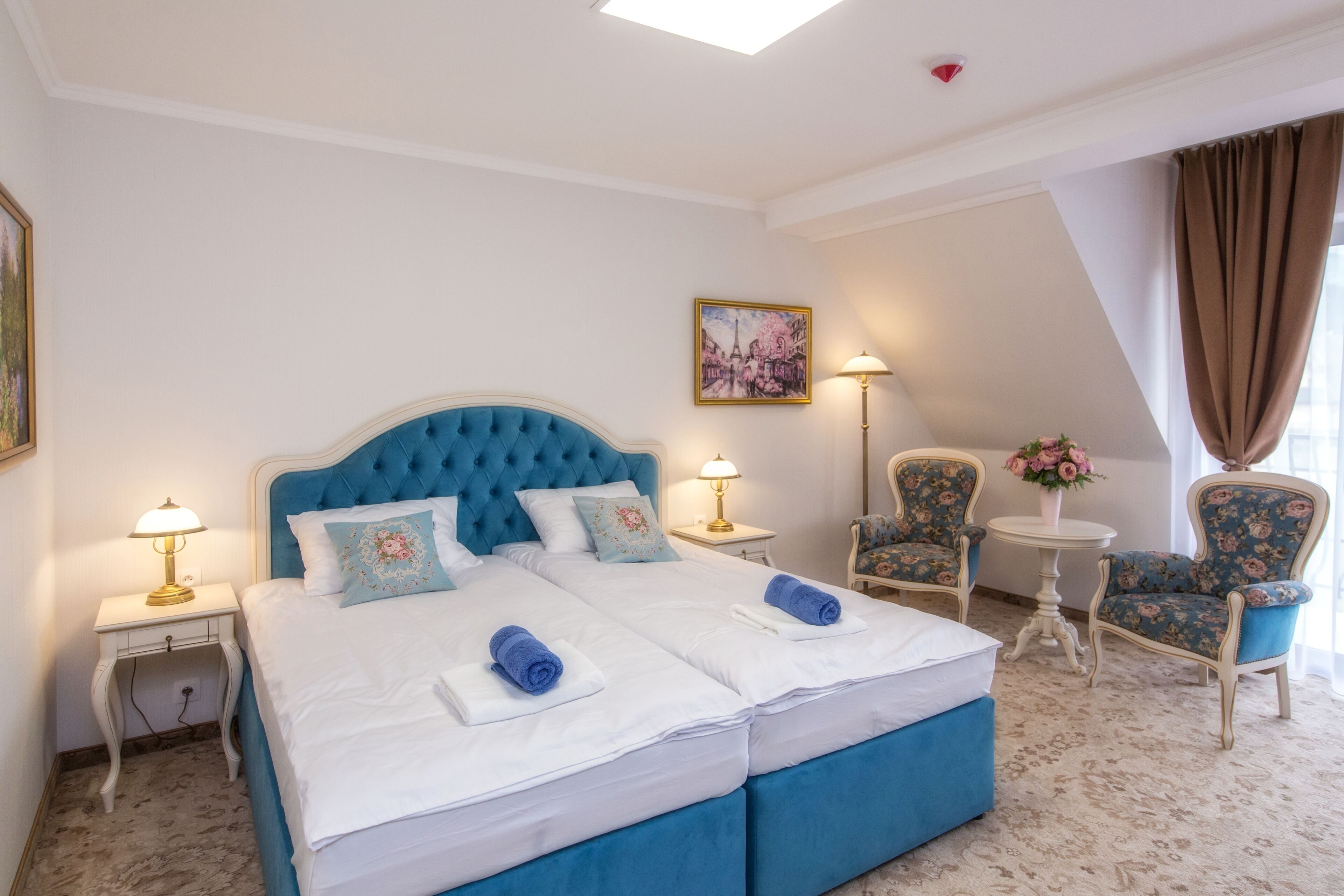 A hotel room with a large bed, two bedside tables, a chair, a small table with a vase, and a painting on the wall.