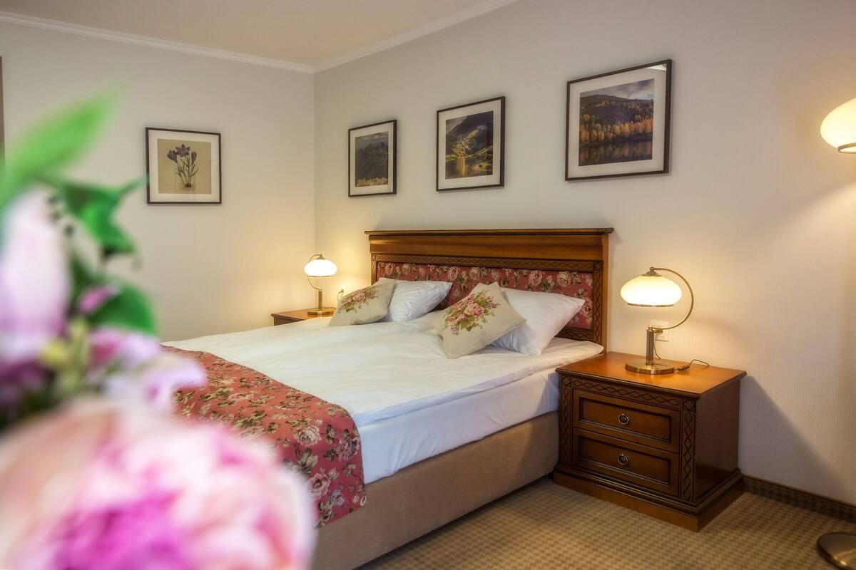 Een net opgemaakt bed met bloemenbedrukking, twee nachtkastjes met lampen, en drie ingelijste foto's aan de muur.