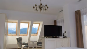 Une chambre d’hôtel comprenant un lit, une télévision, un bureau, deux chaises, un lustre et des fenêtres donnant sur la montagne.