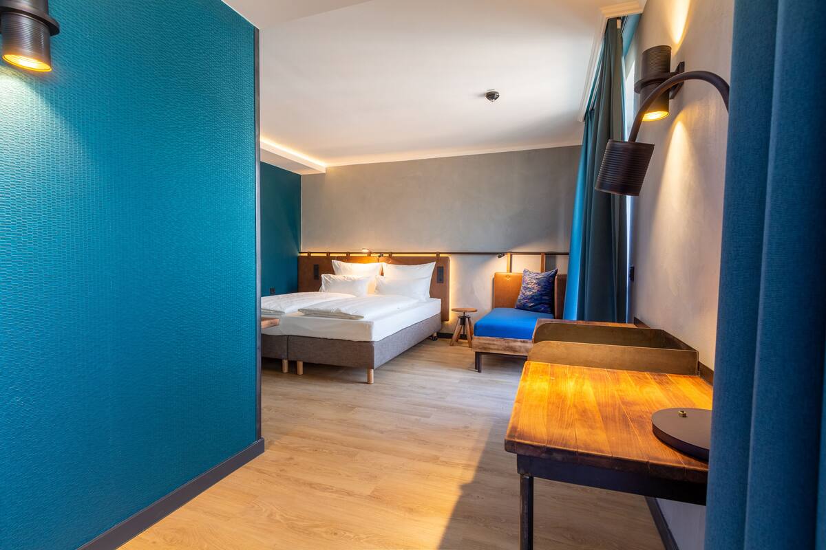 Habitación de hotel con cama, escritorio y silla. Se observa una ventana con cortinas y una lámpara de pared.