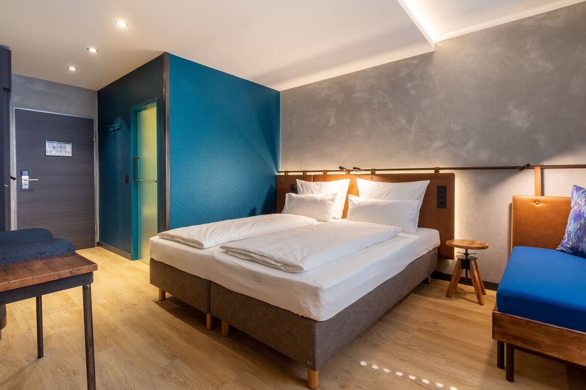 Una habitación de hotel moderna con una cama grande, una mesita pequeña y un sofá azul.