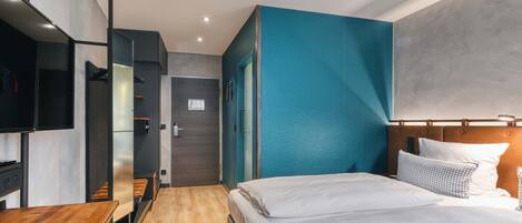 A modern hotel room with a wooden floor, a bed with white linens, a TV mounted on the wall, and a teal accent wall.