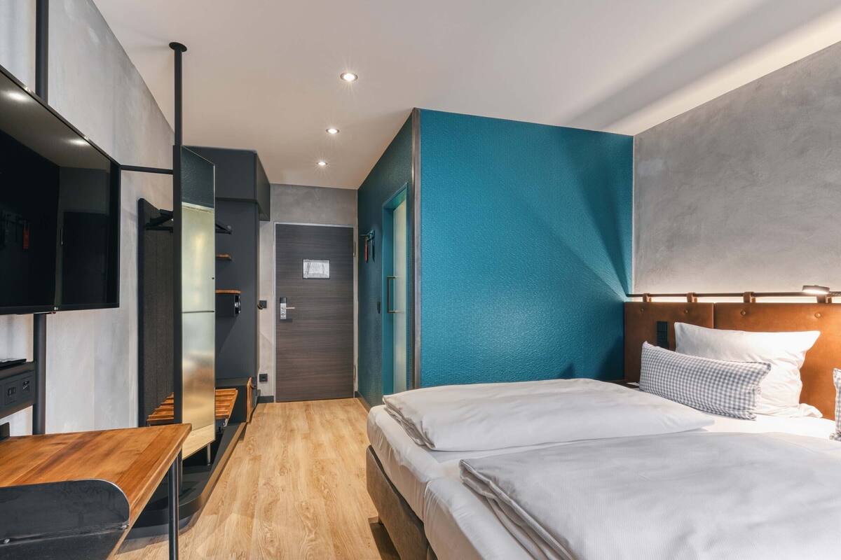 Una habitación de hotel moderna con una cama, un televisor, un banco de madera y un baño visible a través de una puerta de cristal.