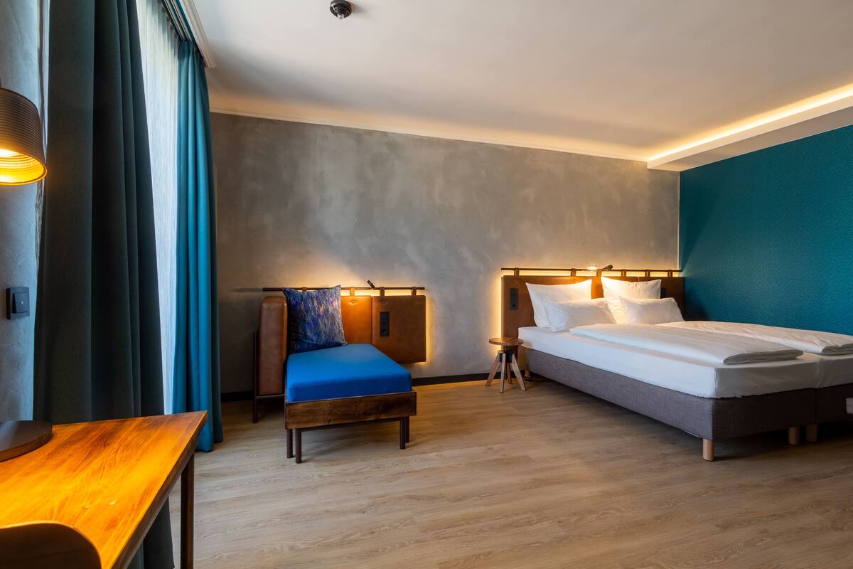 Una habitación de hotel moderna con piso de madera, una cama con ropa blanca, un sofá azul y una pequeña mesa de madera.