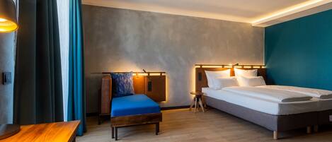 Una habitación de hotel moderna con piso de madera, una cama con ropa blanca, un sofá azul y una pequeña mesa de madera.