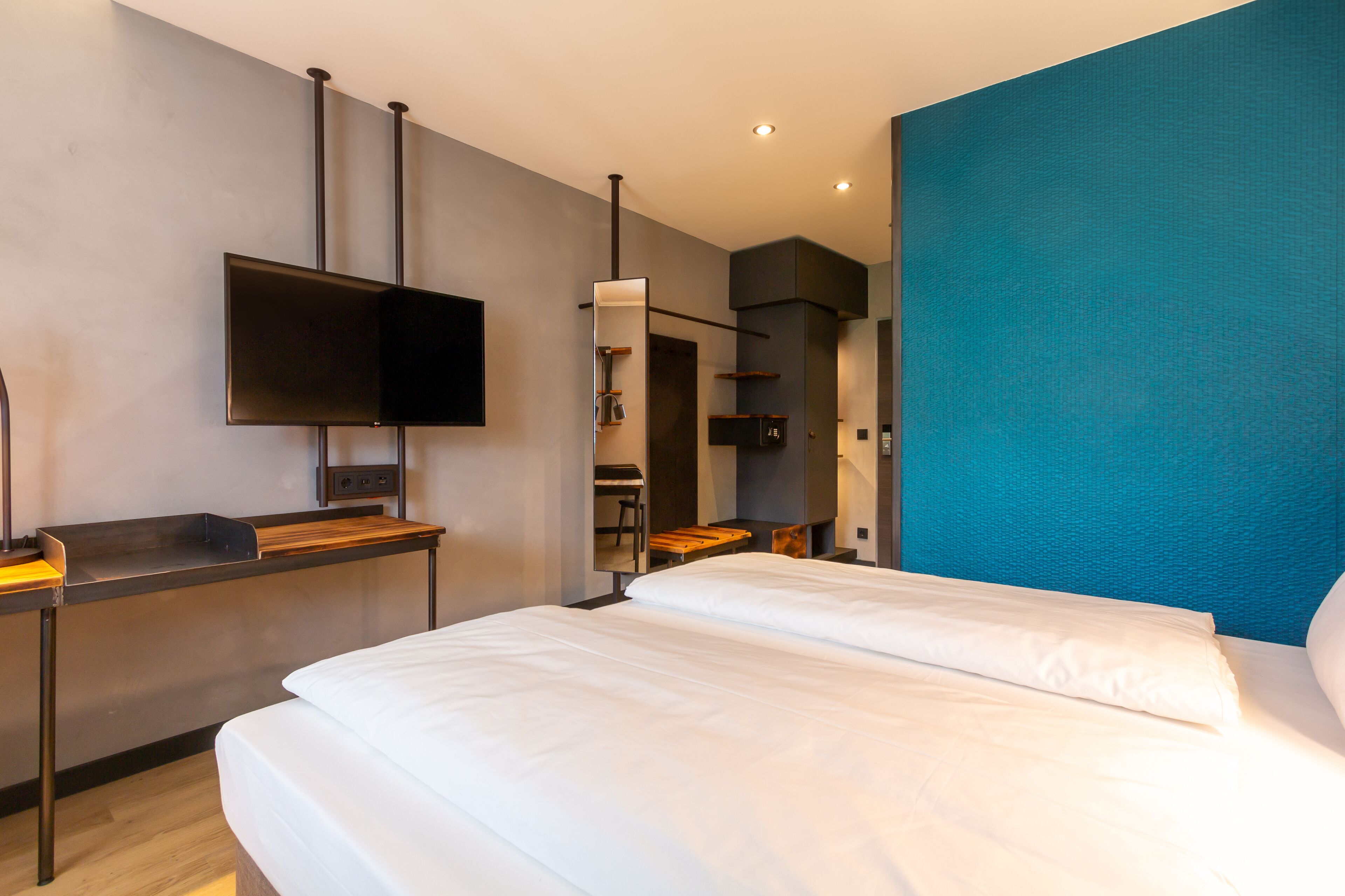 Ein modernes Hotelzimmer mit einem großen Bett, einem flachbildfernseher an der Wand, einem Schreibtisch mit Stuhl und einer blauen Akzentwand.