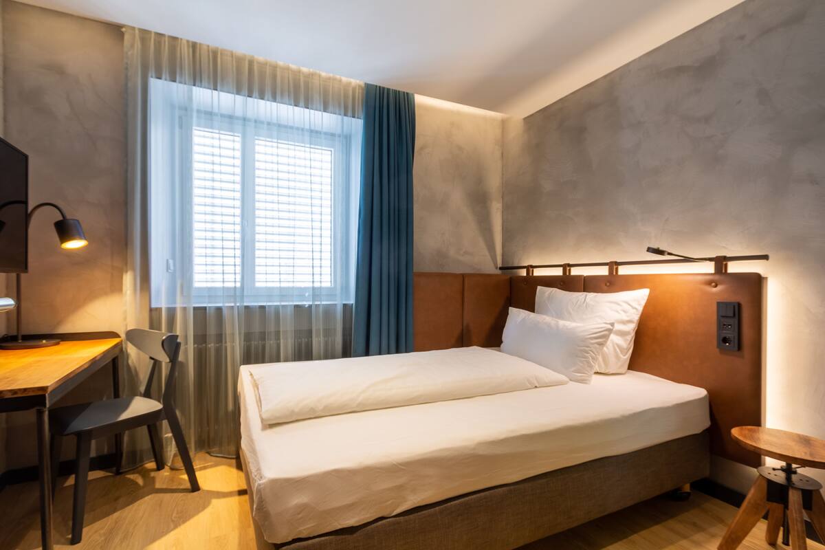 Habitación de hotel con cama, escritorio, silla, ventana con cortinas y cabecera con lámpara.
