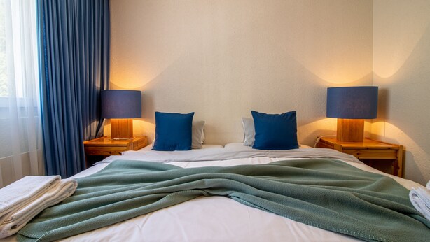 A hotel room with a neatly made bed, two bedside lamps, and a window with blue curtains. - Rothornblick 52 (Arosa)