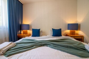 Une chambre d’hôtel avec un lit bien fait, deux lampes de chevet et une fenêtre avec des rideaux bleus.