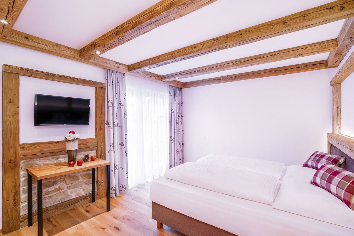 Un dormitorio con techo de madera, chimenea de piedra, televisor de pantalla plana y cama con ropa de cama blanca y almohadas de cuadros.