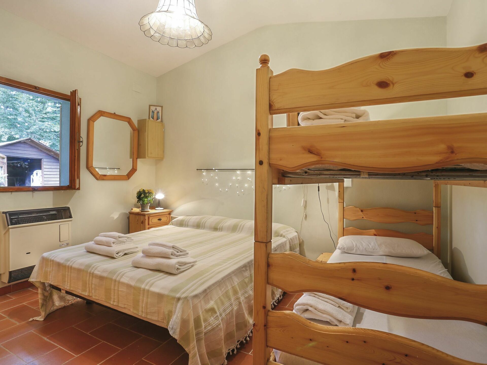 Une chambre avec un lit superposé, une table de chevet avec une lampe, un miroir et une fenêtre donnant sur une maison.