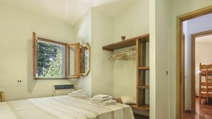 Un dormitorio con una cama, un ventanal, un espejo, un estante de madera con ropa y una puerta que da a otra habitación.