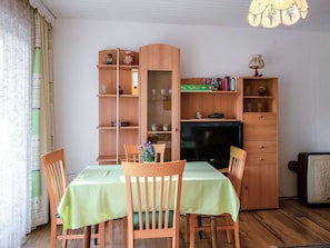 Un coin repas avec une table dressée pour quatre personnes, entourée de chaises en bois. Une bibliothèque contenant divers objets et un téléviseur se trouvent à l’arrière-plan.