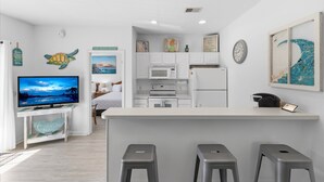 A modern kitchen with white cabinets, a microwave, and a refrigerator. There is a TV on a small table, and a bar area with stools.