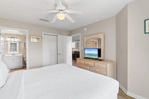 Un dormitorio con cama, ventilador de techo, televisor, cómoda y baño visible a través de una puerta abierta.