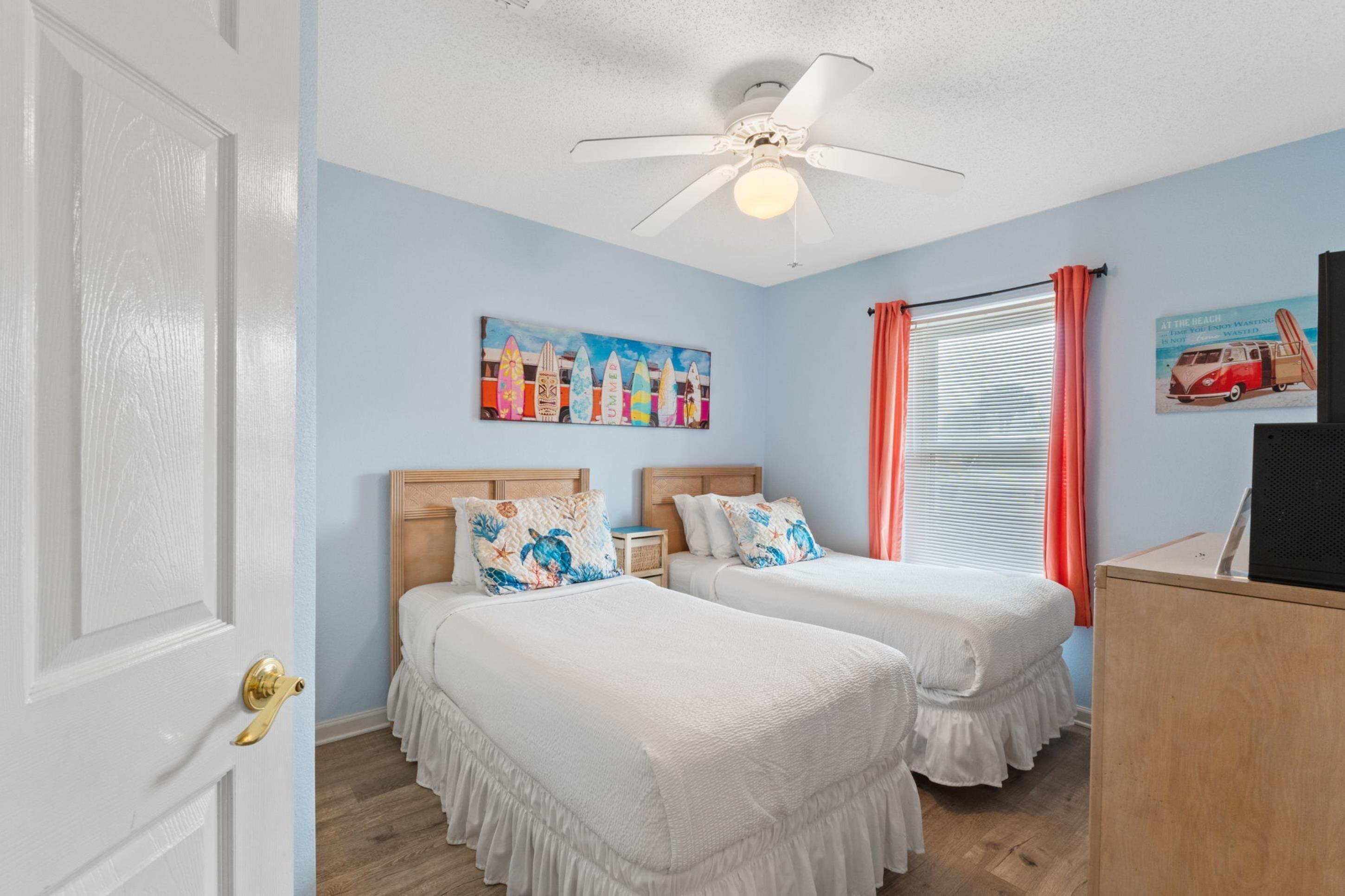 A room with two beds, a ceiling fan, a painting, and a window with curtains.