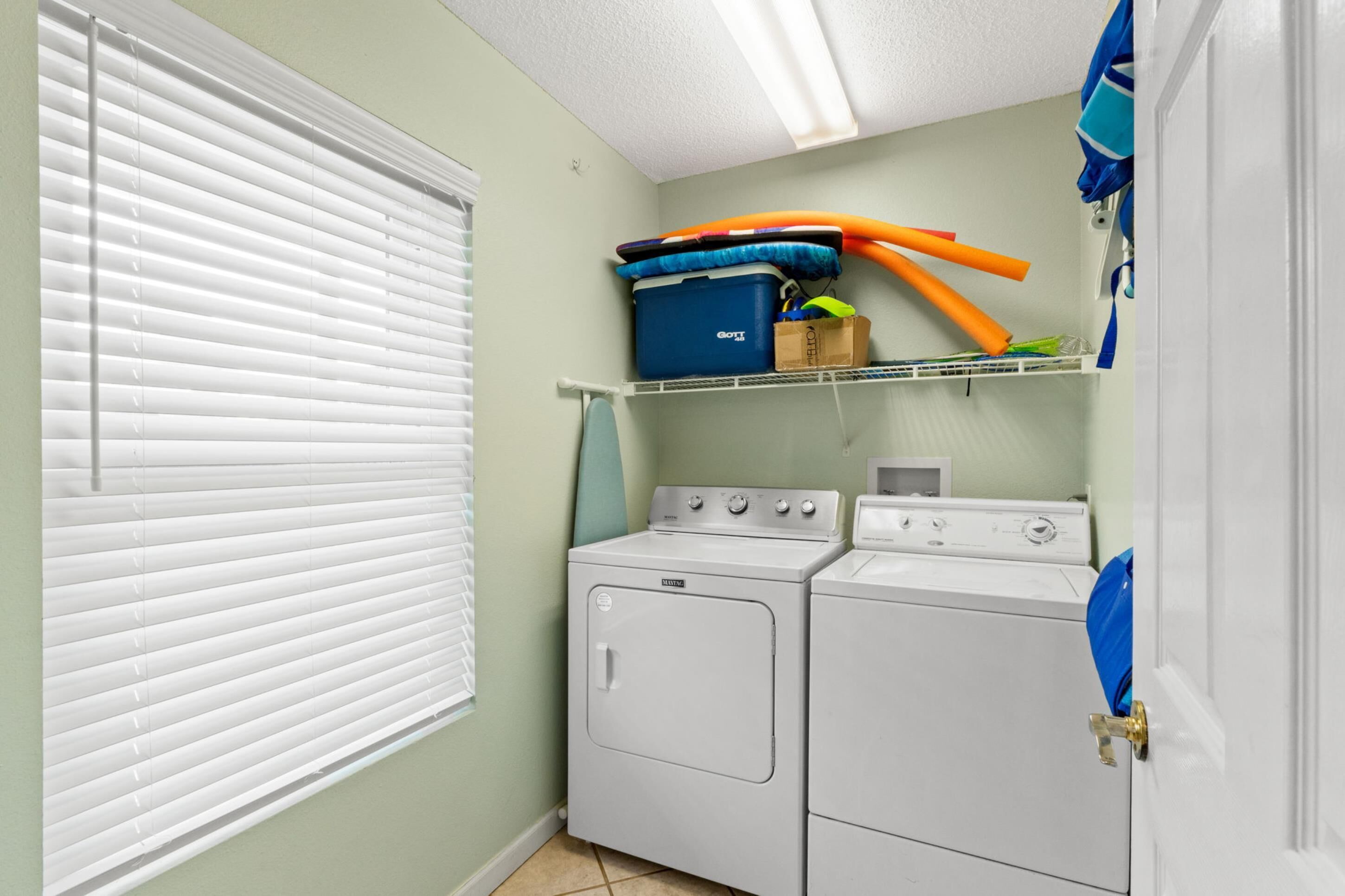 A laundry room with a washer and dryer, a shelf with storage bins, and a window with blinds.