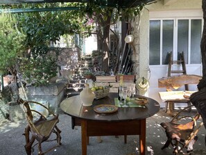 An outdoor patio with a round wooden table, chairs, and a bottle of champagne. There are potted plants and a stone wall in the background.