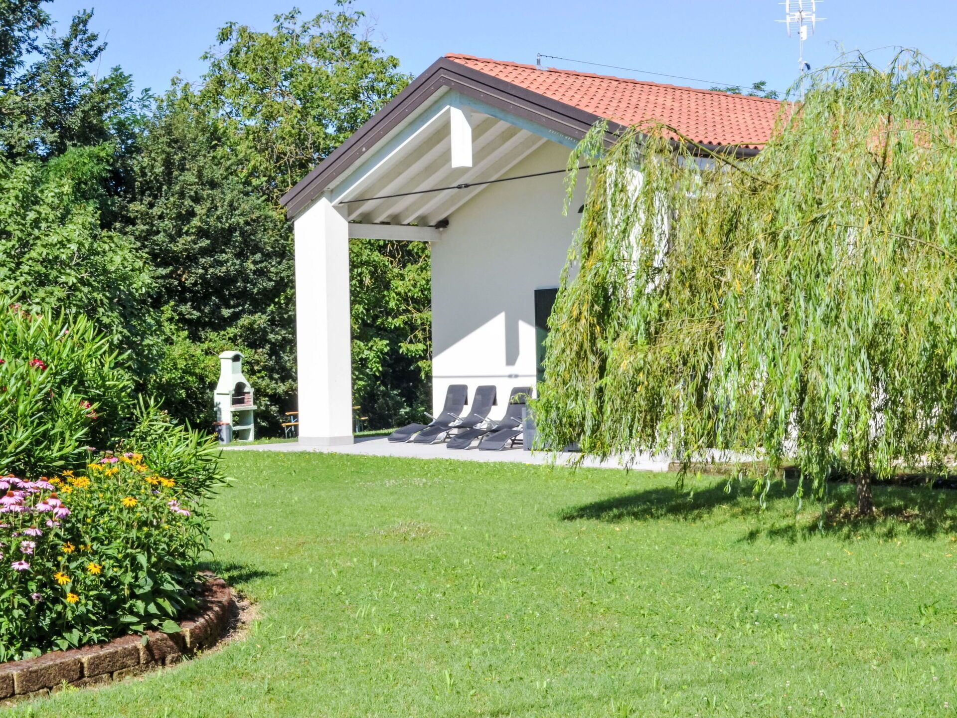 A covered outdoor seating area with a white structure and a red roof, surrounded by lush greenery and flowering plants.