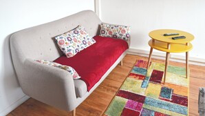 Una habitación acogedora con un sofá gris, almohadones florales de colores, una manta roja y una mesita amarilla con dos controles remotos.