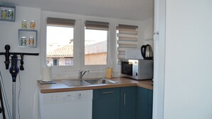 Cocina compacta equipada con lavavajillas, pileta, microondas y tetera. Hay dos ventanas con persianas y un estante pequeño con frascos.