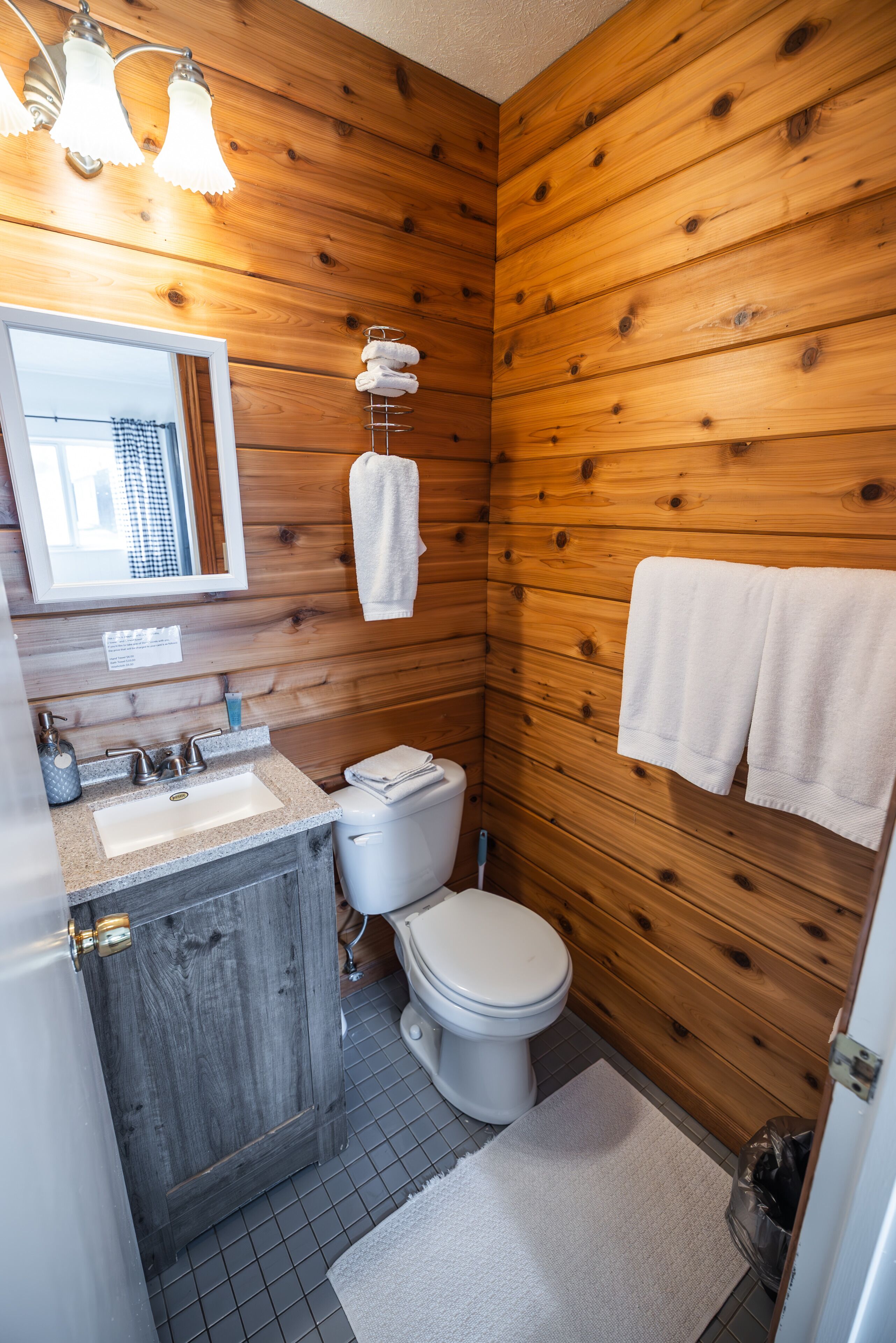 Un baño con paneles de madera, un lavamanos con espejo, un inodoro y toallas colgadas en la pared.
