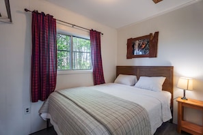 A bedroom with a bed, a nightstand, a lamp, a window with curtains, and a painting on the wall. - Hidden Haven Cottages (Lake City)