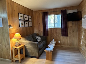 A cozy room with wooden walls, a sofa, a small table with a lamp, a bench, and framed fish pictures on the wall. - Hidden Haven Cottages (Lake City)