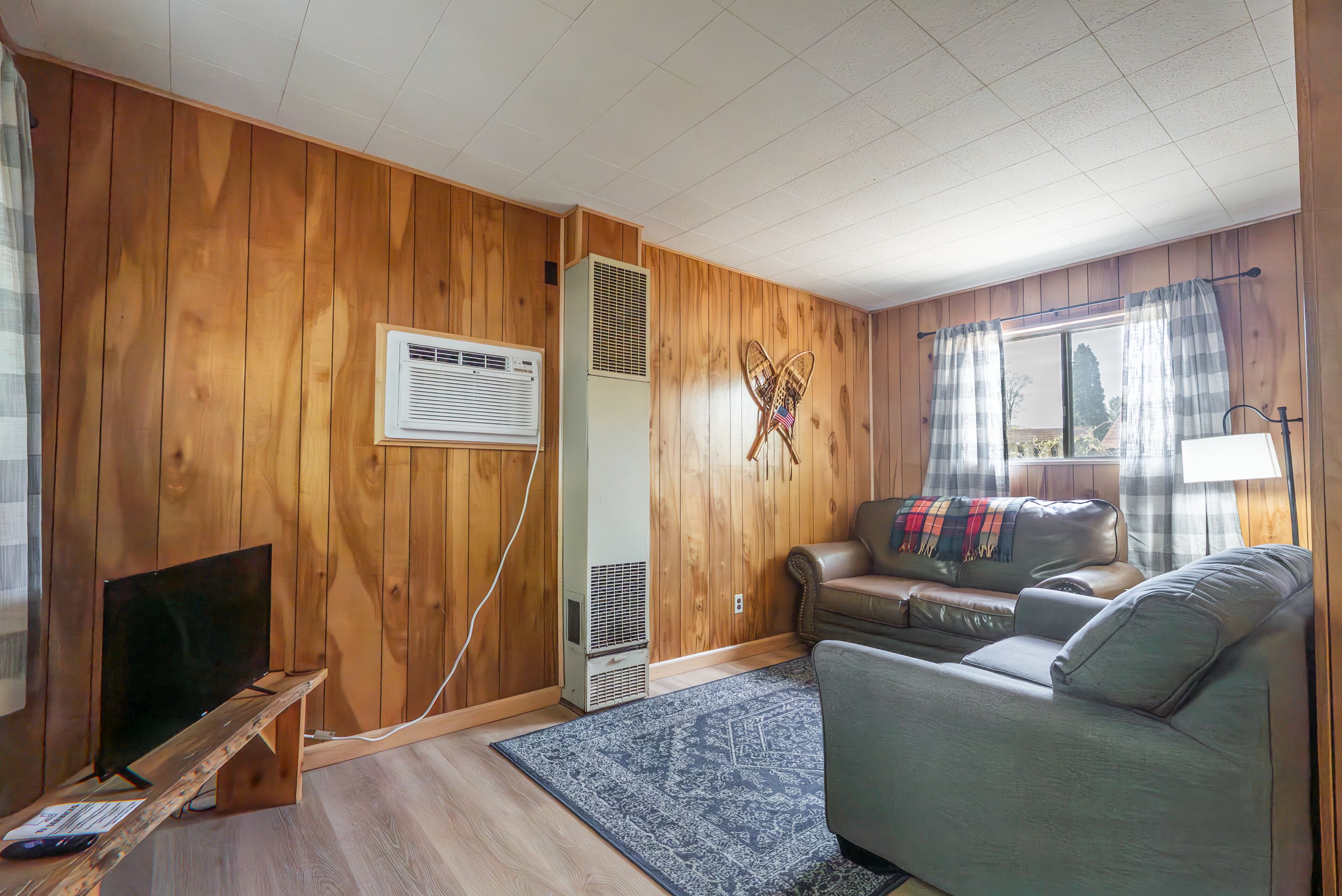 A room with wooden walls, a television, a sofa, a rug, and a window with curtains.
