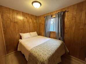 A bedroom with wooden panel walls, a bed with white linens, and checkered curtains. - Hidden Haven Cottages (Lake City)
