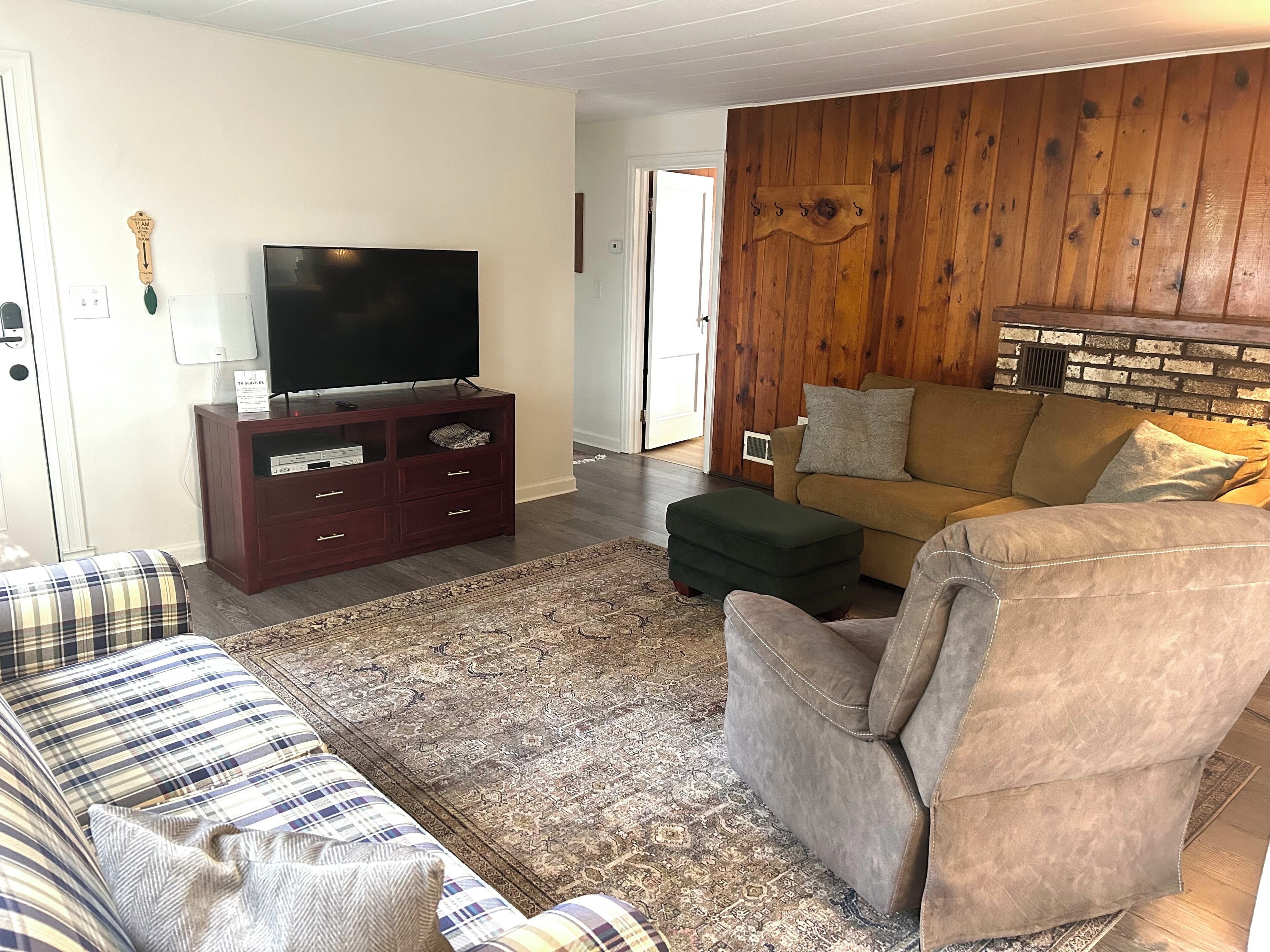 Una sala de estar con un televisor de pantalla plana sobre un mueble de madera, un sofá marrón, un tapete estampado y un panel de pared de madera.