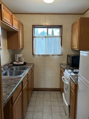 A compact kitchen with wooden cabinets, a window with sheer curtains, a gas stove, a microwave, and a coffee maker. - Hidden Haven Cottages (Lake City)