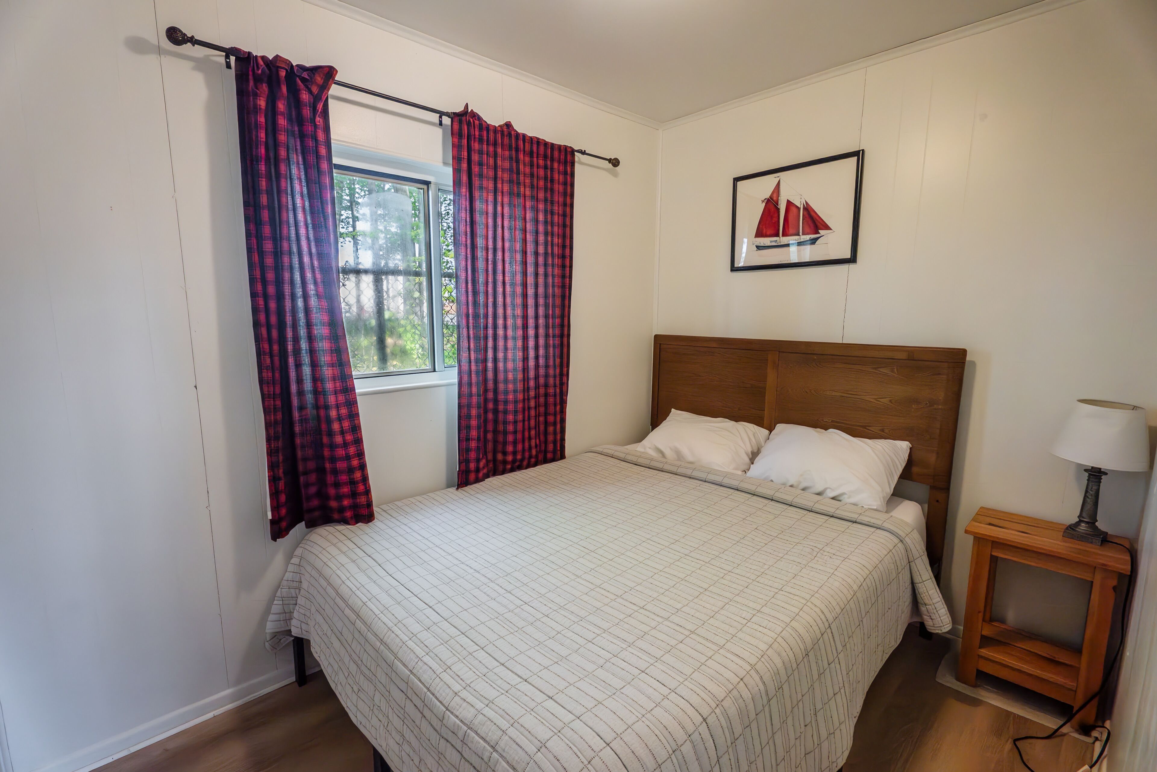 A bedroom with a bed, a nightstand, a lamp, a window with curtains, and a sailboat painting on the wall.