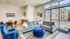 Un salon moderne avec un canapé d’angle gris, des fauteuils bleus et un pouf assorti. On y trouve également un coin repas avec une table et des chaises, et une cuisine en arrière-plan.