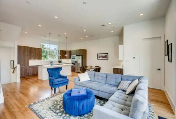A modern living room with a grey sectional sofa, a blue armchair, and a blue ottoman. There is a kitchen in the background with wooden cabinets and a white countertop.