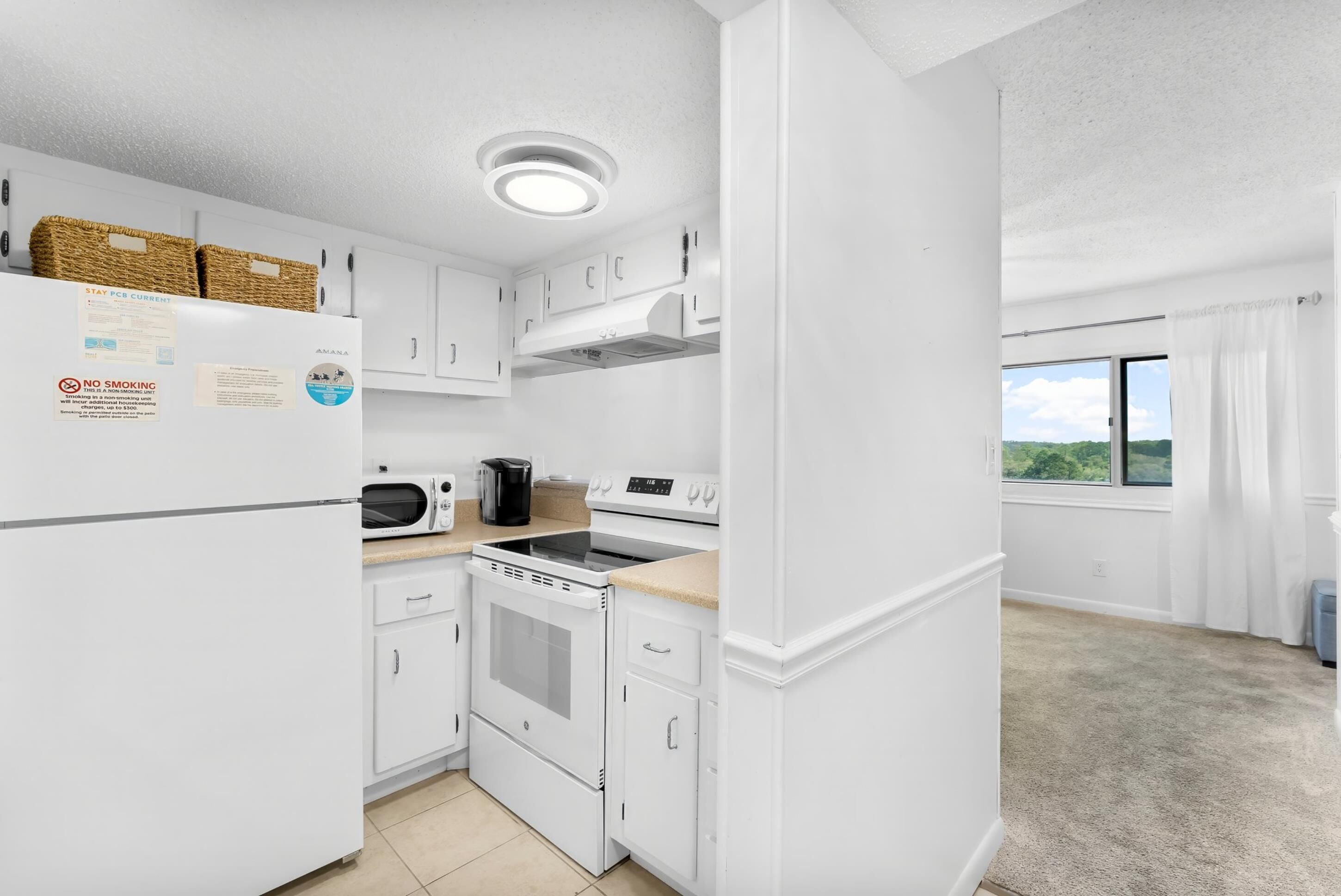 A modern kitchen with white cabinetry, a built-in oven, microwave, and stove. There are baskets on the refrigerator and a window with a view of greenery.