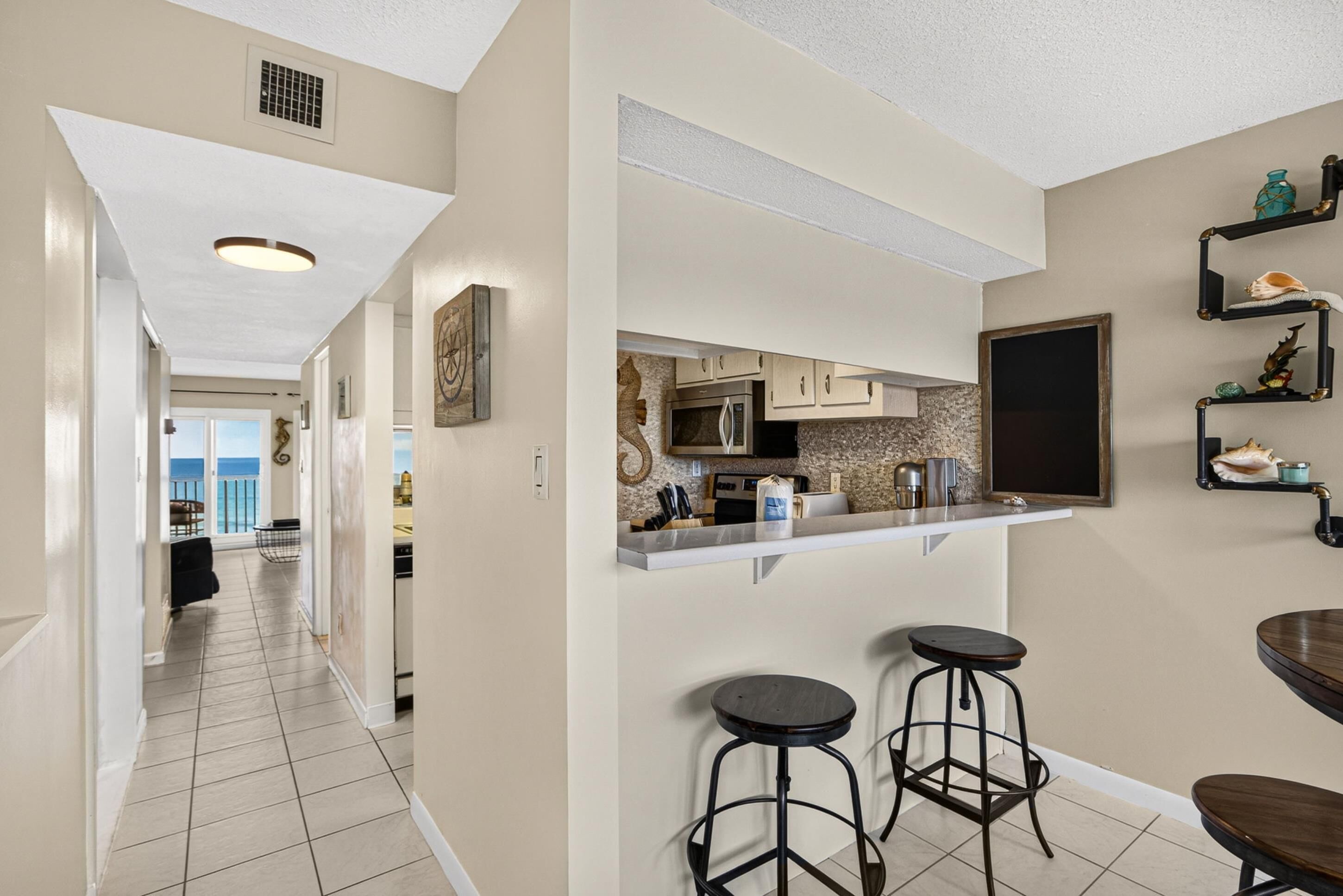 A kitchen with a white countertop, stainless steel appliances, and a large window. There are two bar stools and a wooden table.