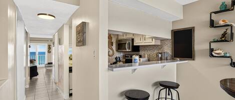 A kitchen with a white countertop, stainless steel appliances, and a large window. There are two bar stools and a wooden table.