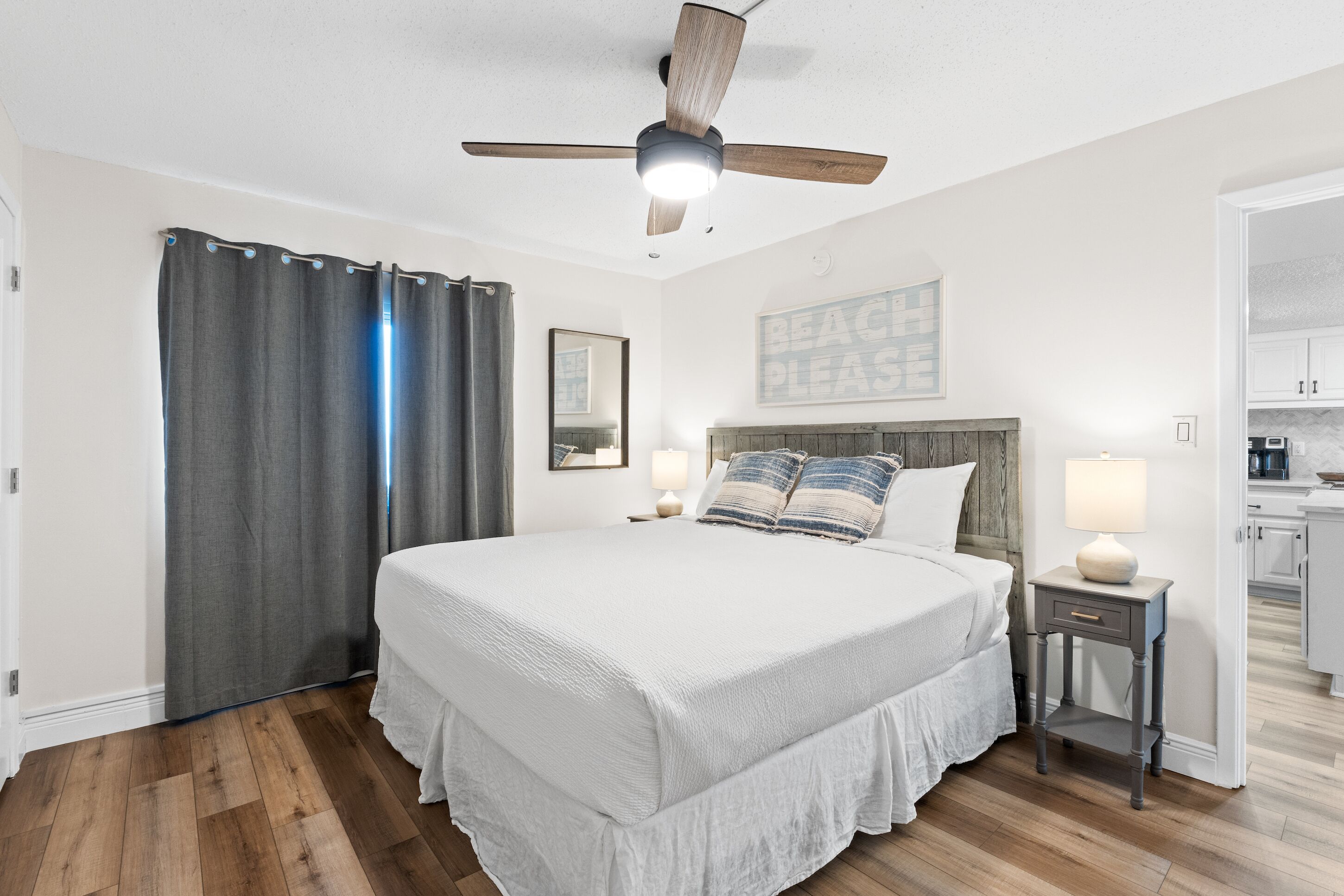 A bedroom with a large bed, a ceiling fan, a nightstand with a lamp, and a kitchen area visible through an open door.