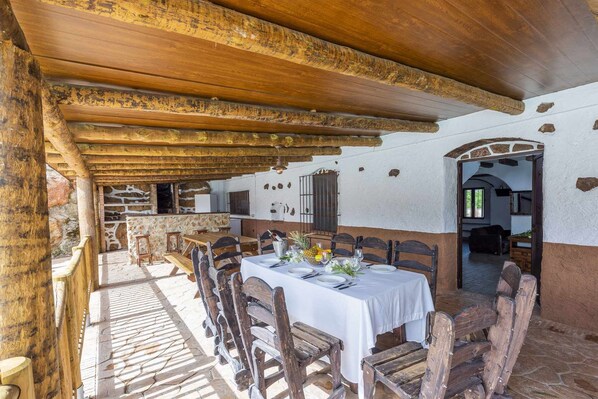 Un comedor al aire libre de estilo rústico con mesas y sillas de madera, un mantel blanco y un muro de piedra de fondo.