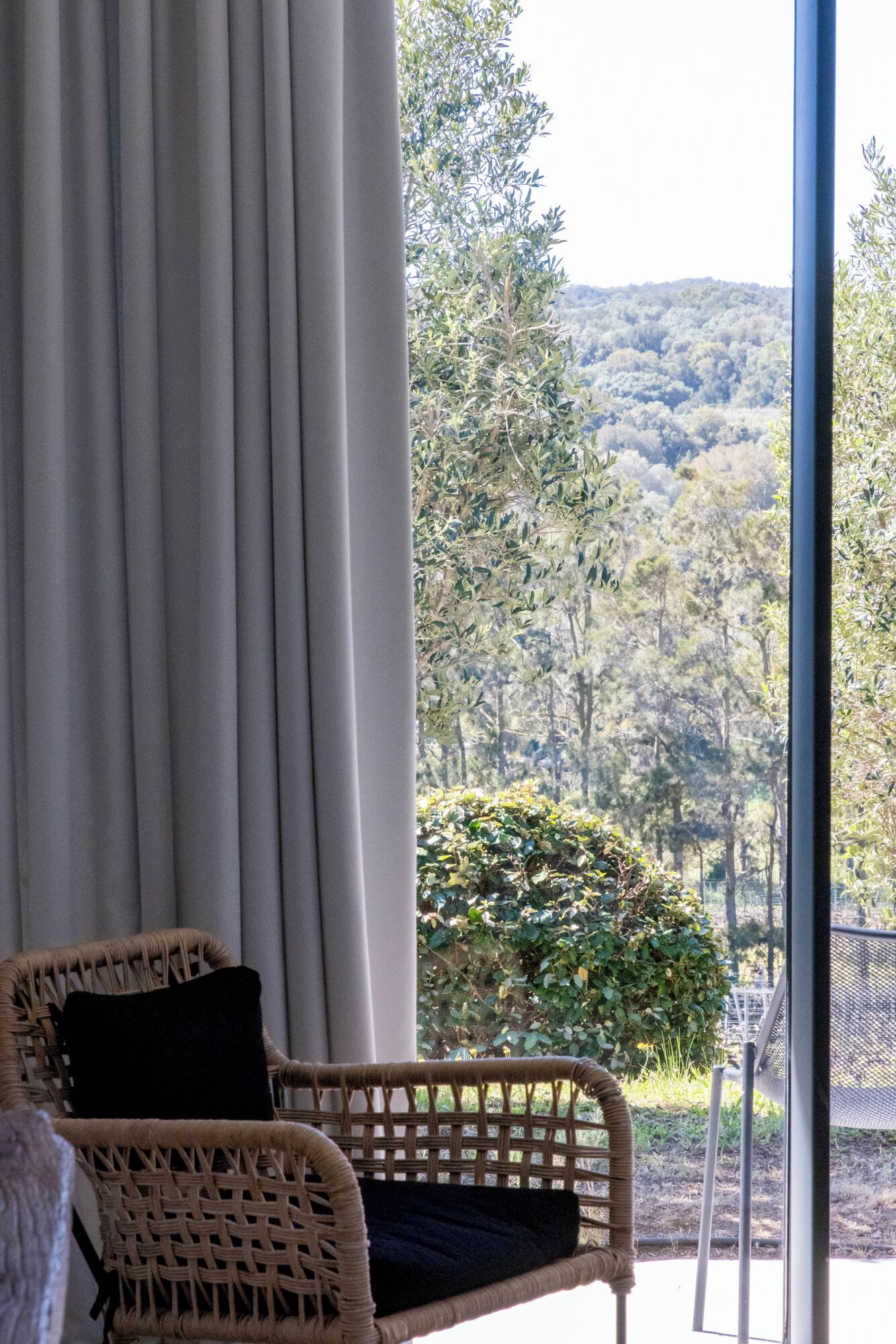 Una silla de mimbre con cojín negro, una ventana con cortinas blancas y vistas a árboles y colinas.