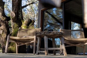 A rustic outdoor seating area with a wooden table and two woven chairs, set against a backdrop of trees and a building with large windows.