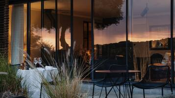 Un área de comedor al aire libre con mesas y sillas, vista al atardecer y un edificio con grandes ventanales.