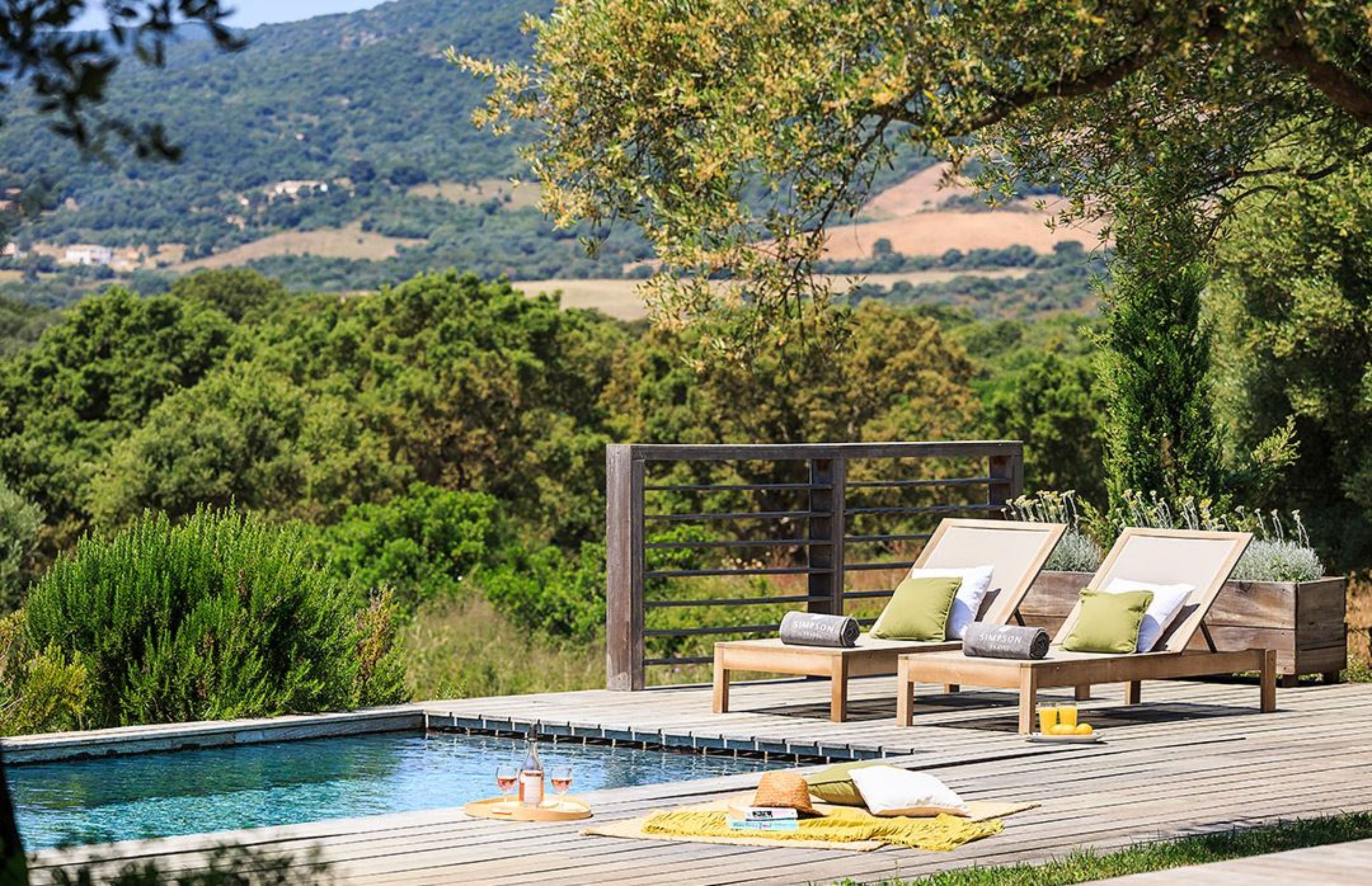 A poolside area with two lounge chairs, a wooden deck, and a view of a hilly landscape.